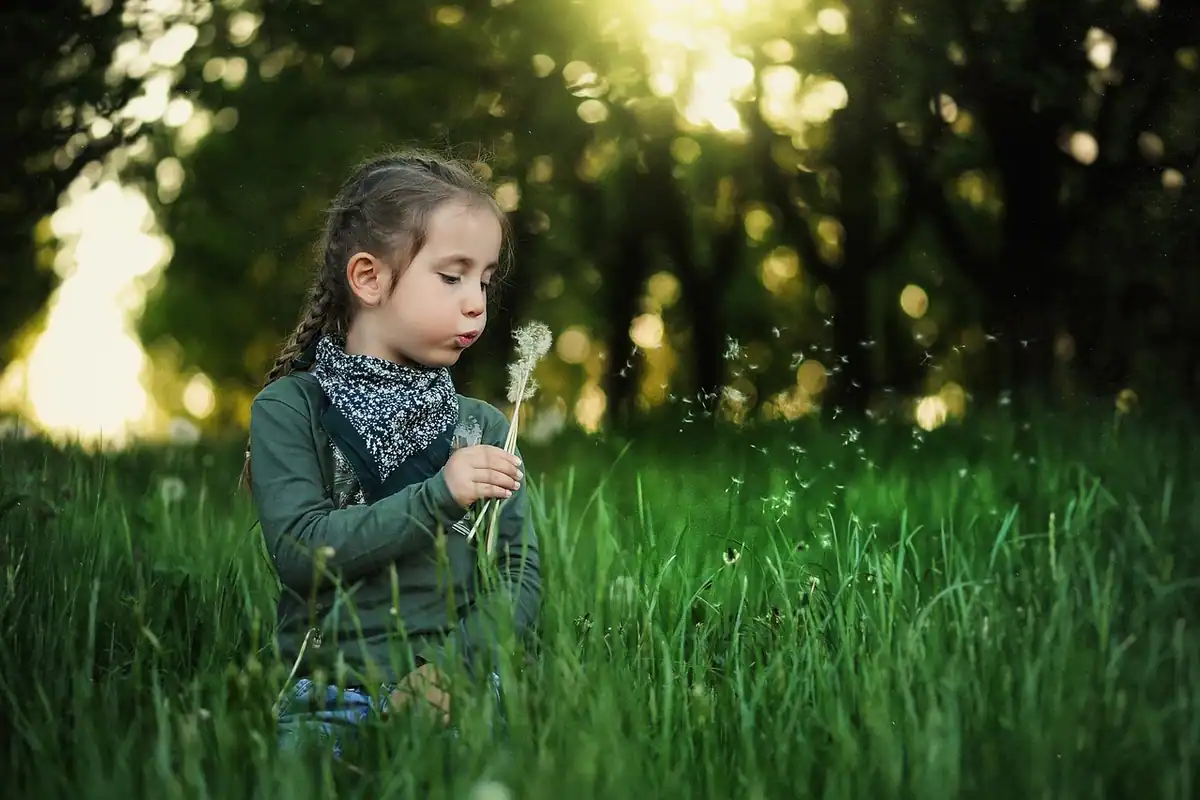 Fetiță într-o pajiște verde suflă un fir de păpădie, în sezonul polenului de primăvară