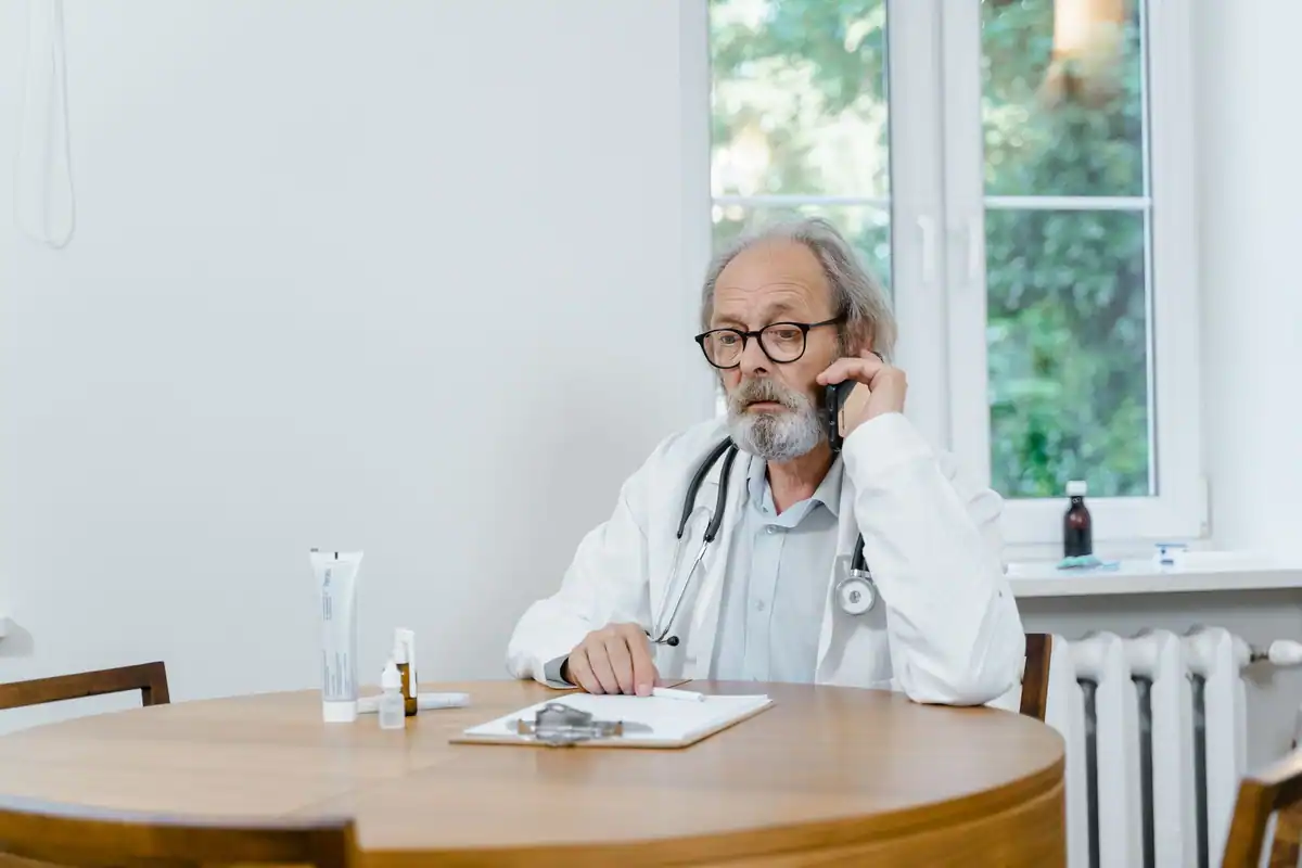 Medic vârstnic cu stetoscop vorbește la telefon la o masă rotundă, într-un cabinet luminos