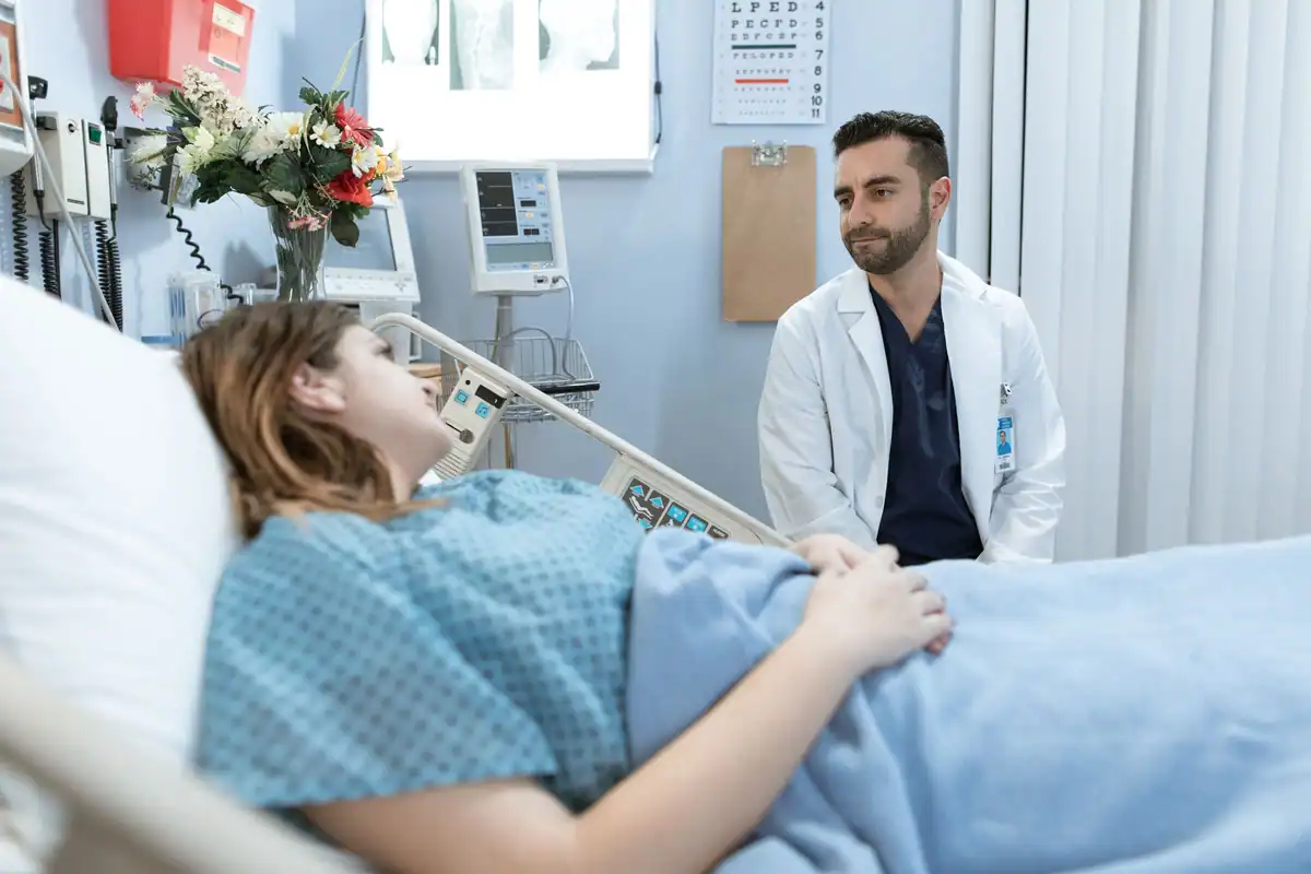 Medic stă lângă o pacientă culcată în pat și o liniștește, într-un salon de spital