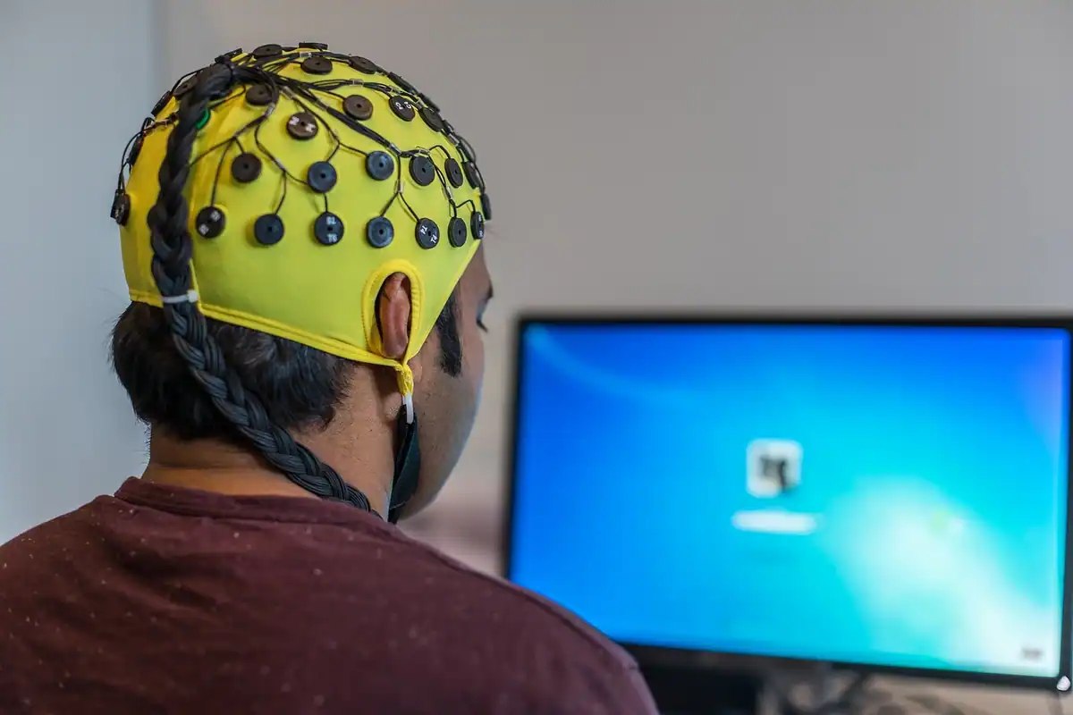 Pacient cu o cască de electrozi galbenă, pentru EEG, în fața monitorului de calculator