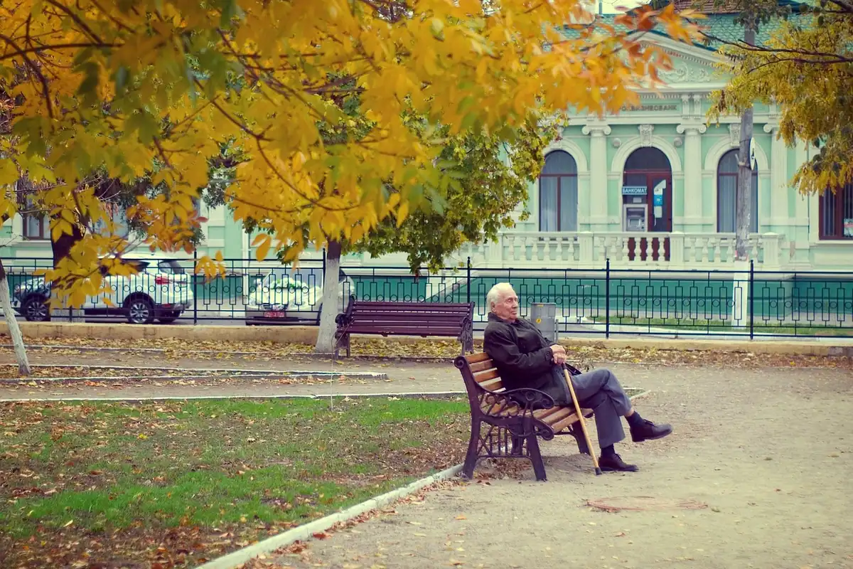Vârstnic stă singur pe o bancă într-un parc, în lumina caldă a toamnei