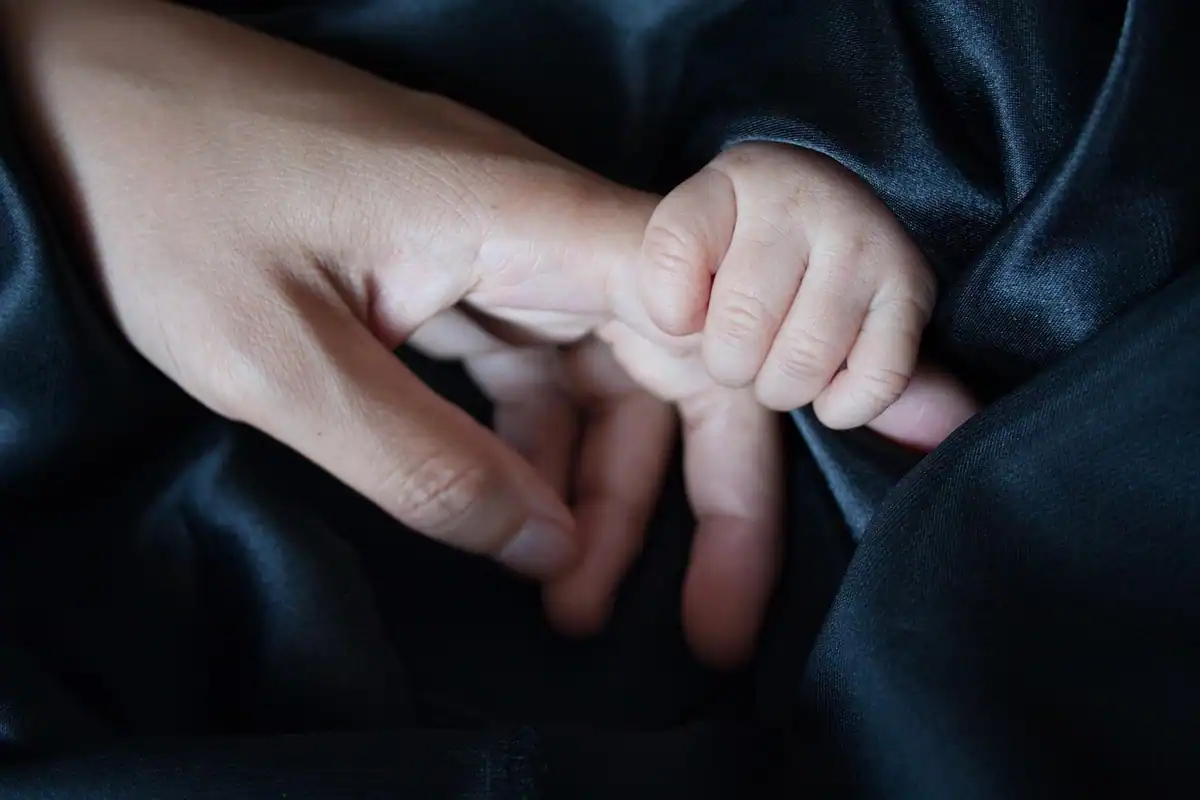 Mâna unui adult ține degetele mici ale unui bebeluș, într-un gest de grijă