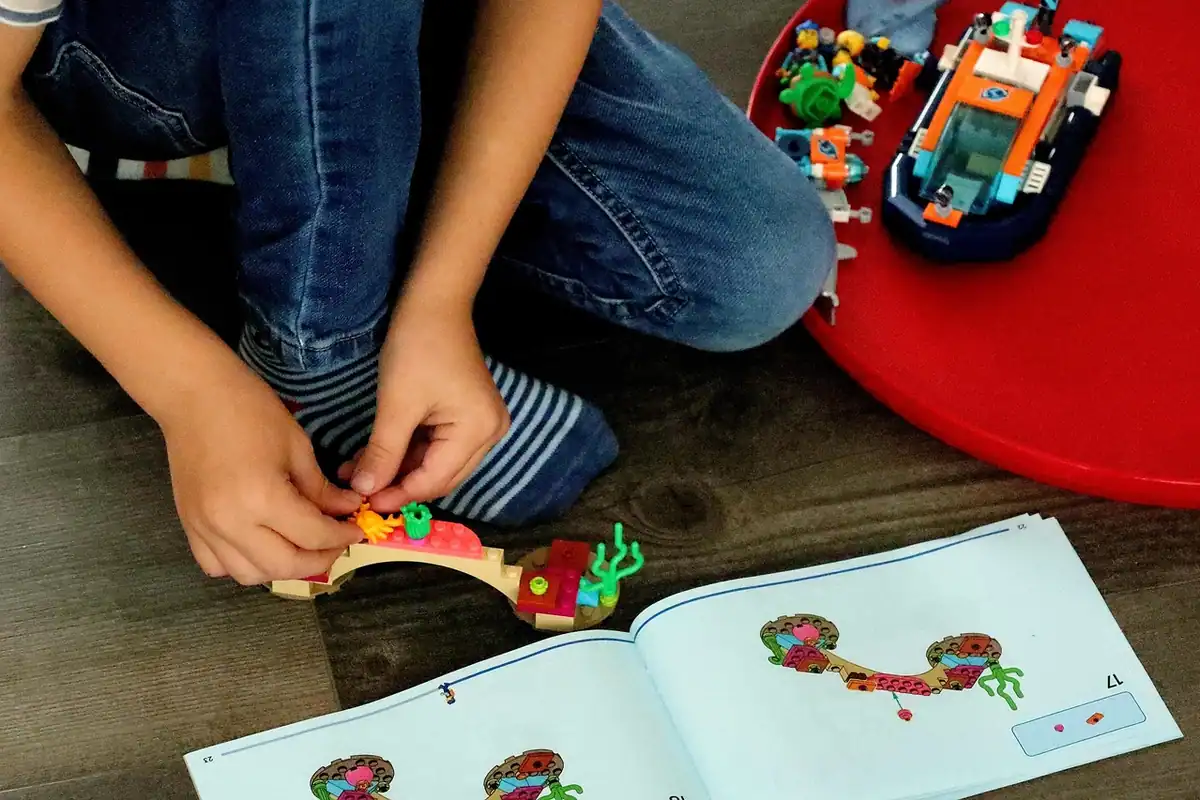 Copil construiește cu piese colorate și lucrează după o carte de instrucțiuni, pe podea